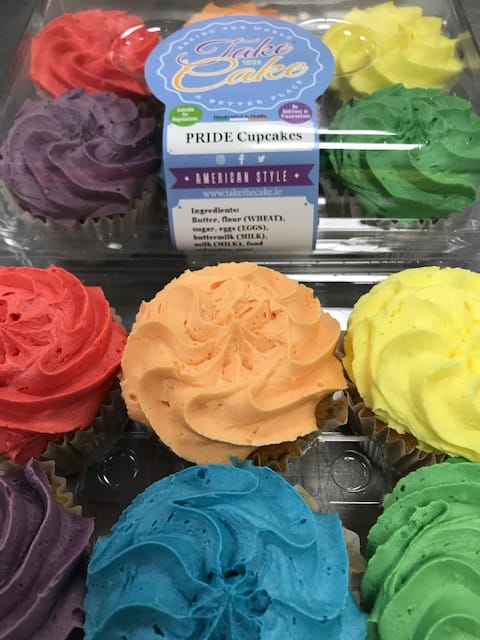 A close-up of a pack of six pride cupcakes with rainbow-colored icing. The packaging label reads 'Take the Cake,' with tags for 'Pride Cupcakes,' 'Suitable for Vegetarians', 'American Style,' and 'Handcrafted in Dublin.' Ingredients are listed at the bottom.